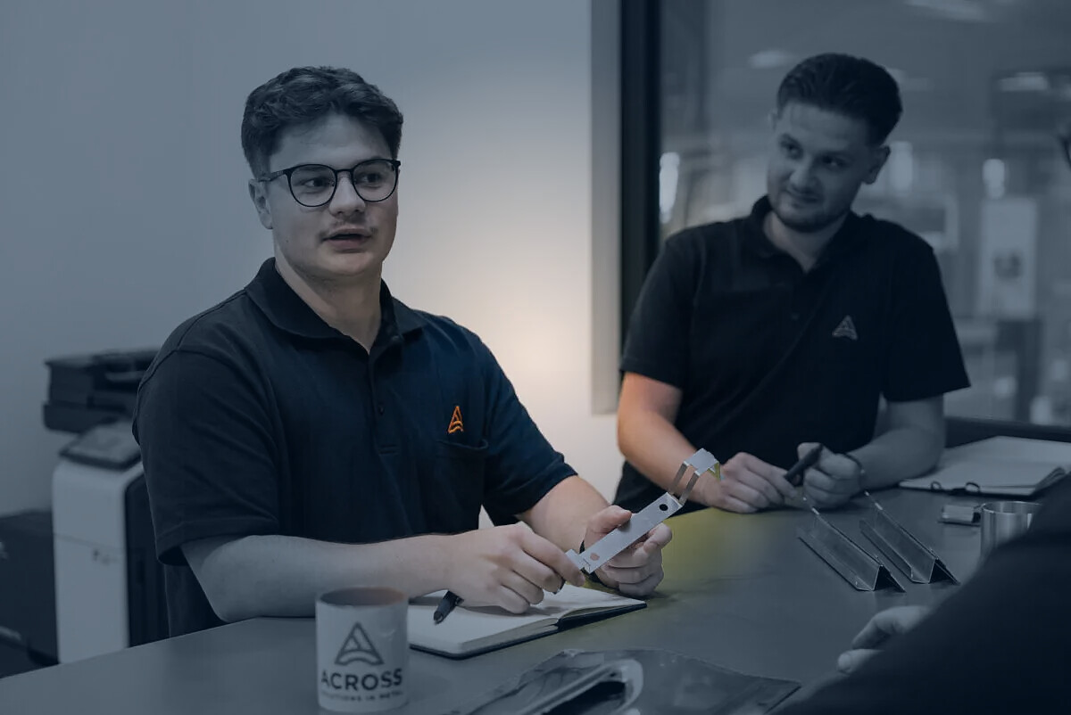 Mitarbeiter von ACROSS Solutions in Metal besprechen ein Metallbauteil.